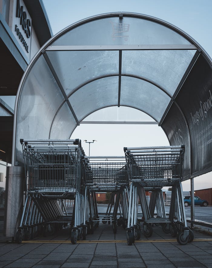 A group of metal shopping carts under a transparent shelter at a supermarket parking lot.